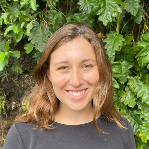 A young woman with long hair smiles at the camera, standing in front of a lush green background featuring leaves and plants.