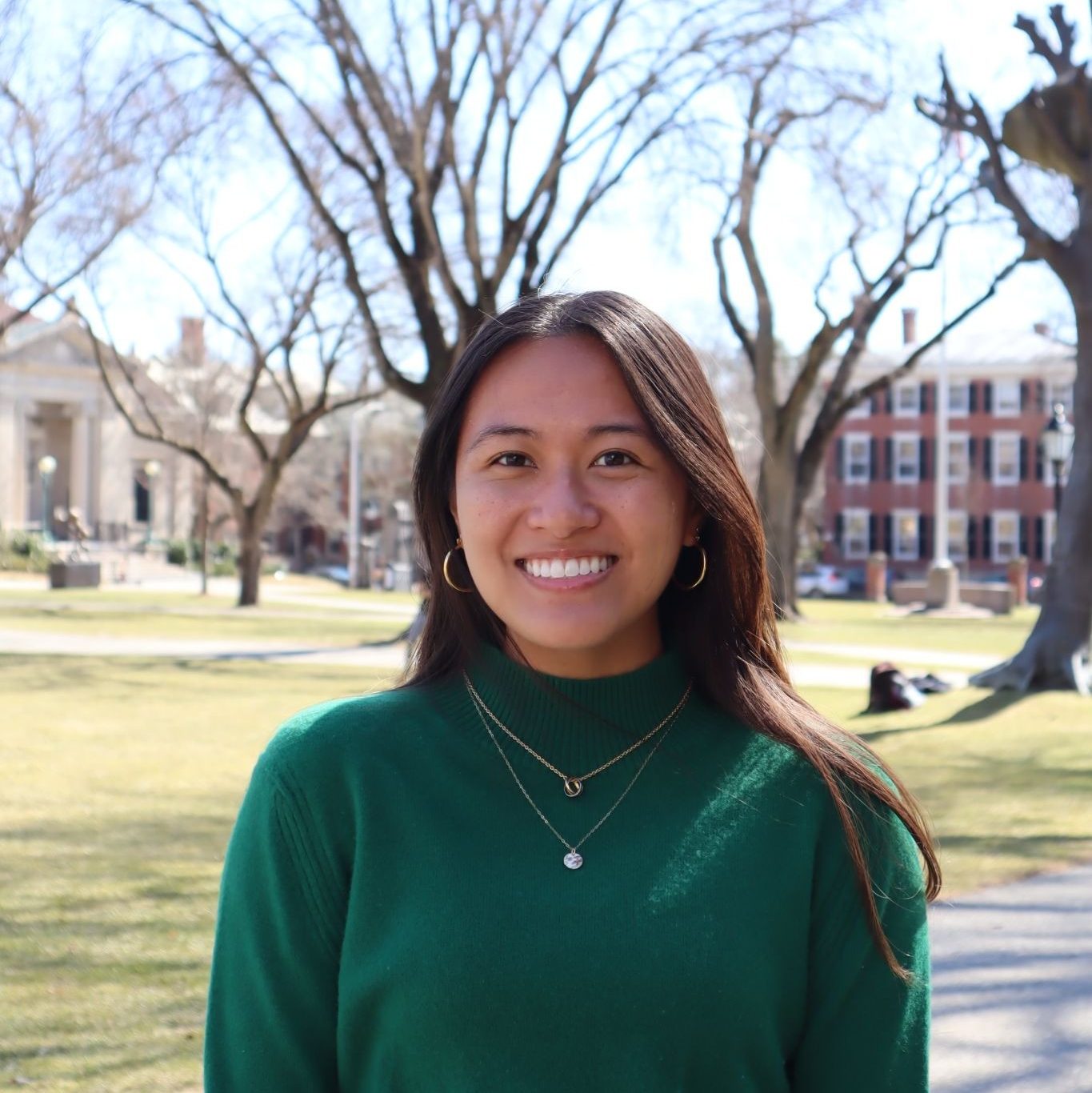 A smiling individual wearing a green sweater stands in a park setting with trees and buildings in the background.