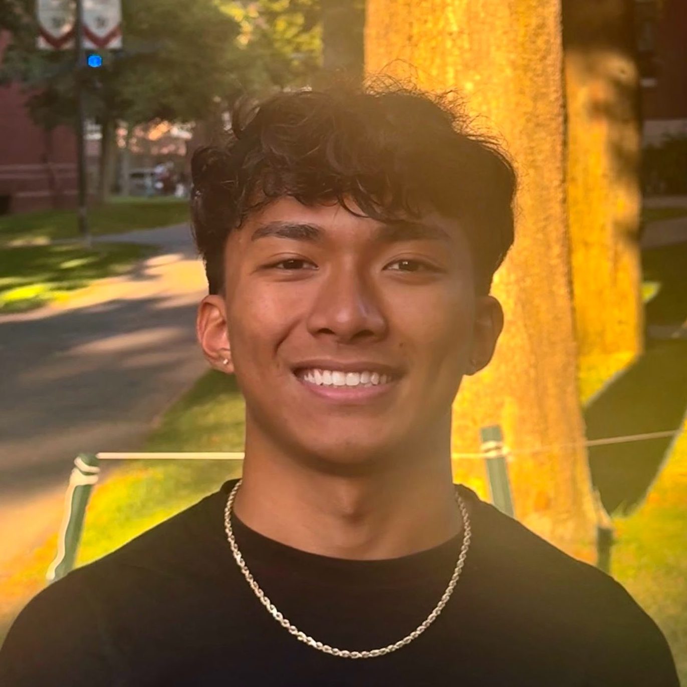 A smiling young man with curly hair wearing a black shirt and a chain necklace, standing outdoors with trees and buildings in the background.